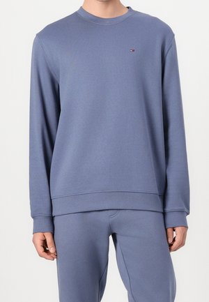 Mężczyzna ubrany w niebieską bluzę z długim rękawem i pasujące spodnie z małym logo Tommy Hilfiger na piersi.