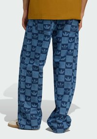 Blue denim pants featuring a checkerboard pattern with dark blue Adidas logos. They have a relaxed fit and are styled with beige shoes.
