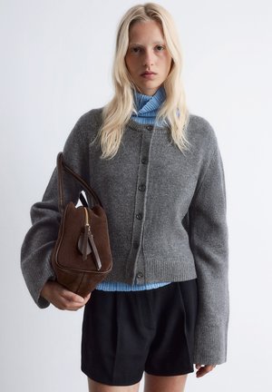 Grau gestrickte Weste mit Rundhalsausschnitt und Knopfleiste, getragen über einem hellblauen, gerippten Rollkragenpullover, kombiniert mit schwarzen Shorts und einer braunen Tasche.
