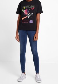 Camiseta de algodón negra con un gráfico colorido de un cohete y planetas. Combinada con jeans skinny azules y zapatillas grises.