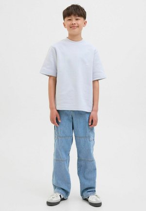 Světle šedé oversized tričko s krátkými rukávy, v kombinaci se světle modrými volnými džíny s cargo kapsami. Bílé tenisky s černými akcenty.
