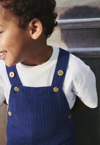 Blue corduroy overalls with four wooden buttons, worn over a white crew-neck t-shirt. Texture visible in fabric's ridges.