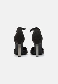 Chaussures à talons hauts en daim noir avec un talon bloc métallique. Présentent une sangle de cheville à l'arrière et des détails en mesh sur les côtés.