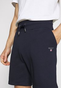 Homme portant un short GANT bleu marine avec taille à cordon et poches latérales, associé à un t-shirt blanc.