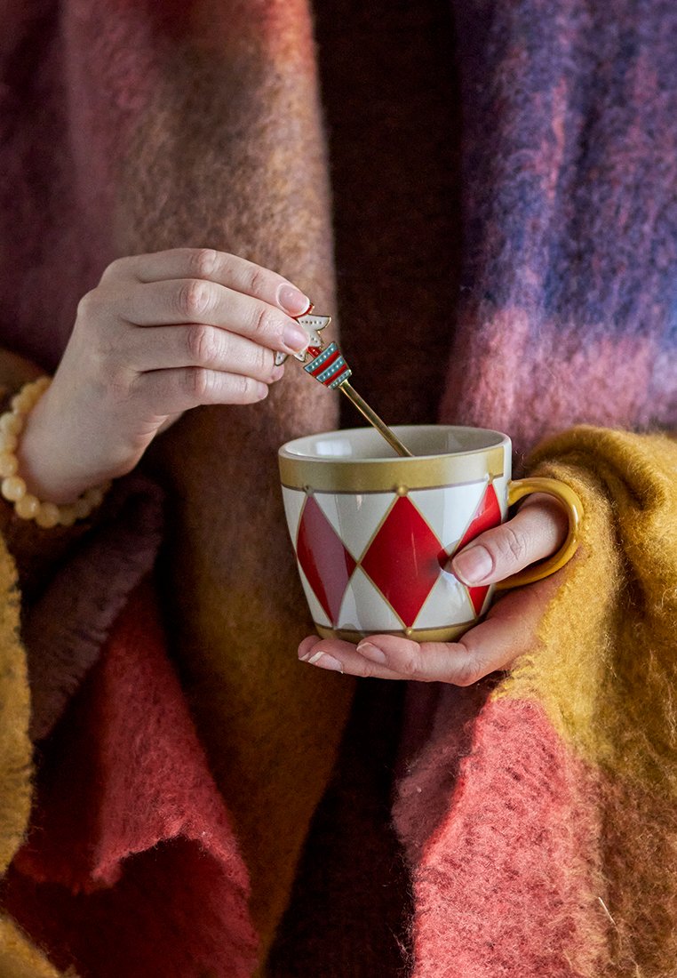 Keramiktasse mit rotem Diamantmuster und goldenem Rand, gehalten von einer Person, die einen braunen und goldenen gestrickten Schal trägt und mit einem goldenen Löffel umrührt.
