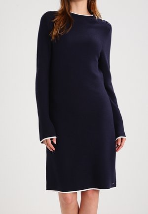 Kobieta ubrana w granatową, dzianinową sukienkę z długim rękawem sięgającą do kolan, z białym obszyciem na mankietach i dole oraz ozdobnymi guzikami na jednym ramieniu.
