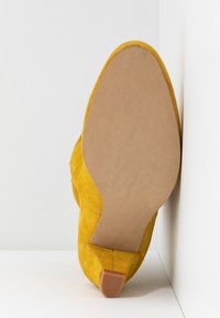 Semelle de chaussure en daim jaune face vers l'avant avec un petit talon en caoutchouc, positionnée dans un angle entre un mur blanc et le sol.