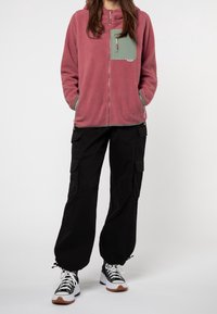 Person iført en mauve fleecejakke med grøn brystlomme, sorte cargo-bukser og sort-hvide platform sneakers, der står foran en hvid baggrund.