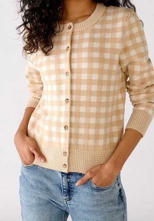 Femme portant un cardigan boutonné à carreaux beige et blanc avec poignets et ourlet côtelés, associé à un jean denim bleu clair, mains dans les poches.