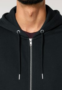 Fekete, cipzáras kapucnis felső, pamutból készült, ezüst cipzárral és fém szemekkel, zsinórokkal és puha, texturált anyaggal.