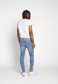 Blå denim skinny jeans med en läder Lee-logotyp på bakre midjan, ihop med en grå t-shirt och vita sneakers.