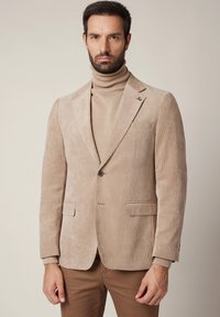 Mężczyzna z ciemnymi włosami i brodą, ubrany w beżową marynarkę z kordurowego materiału, dopasowany beżowy golf oraz brązowe spodnie, stojący na jednolitym tle.