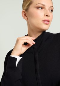 Zwarte zip-up trui met hoge kraag, geribbelde textuur en witte shirtmanchetten die uitkomen. Glad materiaal met een slim-fit vorm.