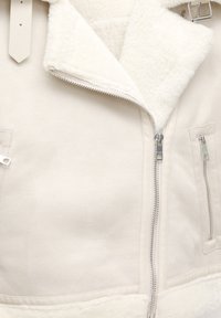 Biała kurtka z sztucznego kożuszka z zamkiem błyskawicznym z przodu, kołnierzem i dwoma bocznymi kieszeniami. Teksturowana powierzchnia z gładkimi akcentami i srebrnymi elementami.