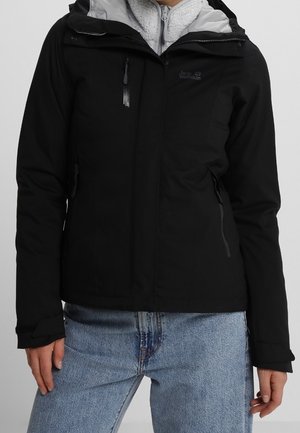 Persona che indossa una giacca nera con zip e cappuccio e fodera interna in pile grigio chiaro, abbinata a jeans azzurri.