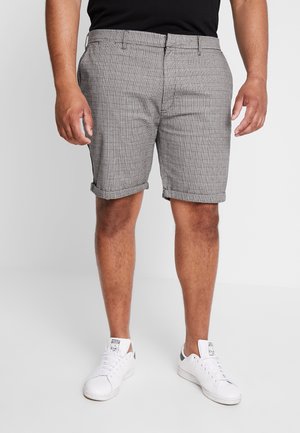 Mand iført grå ternede shorts med opsmøgede kanter og hvide sneakers, stående mod en ensartet lys baggrund.