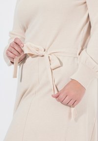 Abito in maglia beige con cintura in vita, dotato di maniche lunghe e orlo curvo. Il tessuto ha una finitura morbida e testurizzata.
