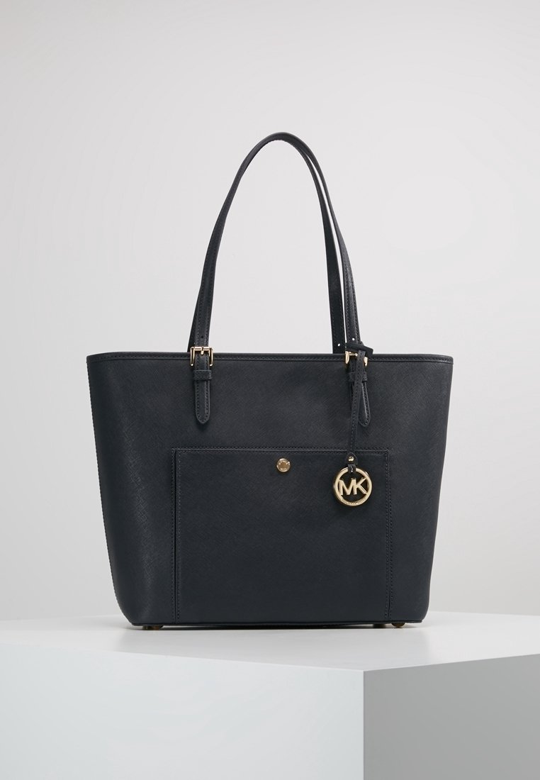 Sac cabas en cuir noir avec doubles bretelles, poche avant et breloque dorée circulaire avec logo MK suspendue à une bretelle.