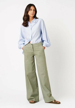 Frau trägt eine hellblaue Nadelstreifenbluse, weite olivgrüne Hose und Sandalen, steht mit den Händen in den Taschen vor weißem Hintergrund.