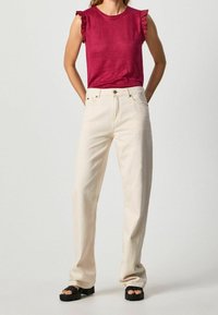 Žena v bezramenném červeném topu s volány na ramenou, v kalhotách s vysokým pasem krémové barvy a černých platformových sandálech, stojící s rukama za zády.