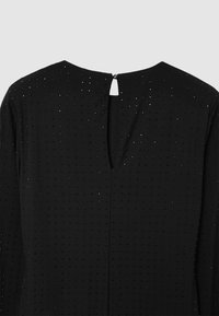 Schwarzes Kleidungsstück mit einem strukturierten Muster aus kleinen Punkten. Der Rücken hat eine Schlüssellochöffnung, die mit einem Knopf gesichert ist. Glatte Stoffoberfläche.