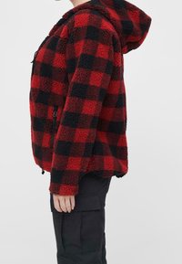 Czerwono-czarna kurtka polarowa w kratę z kapturem, wyposażona w boczną kieszeń na zamek i teksturowaną, puszystą tkaninę.