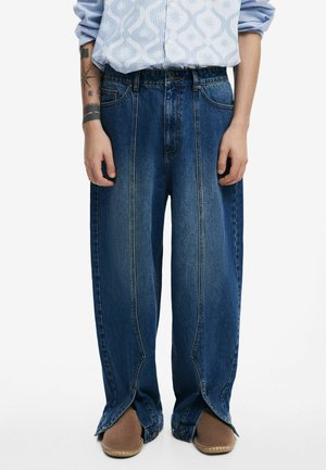 Wide-leg blue denim trousers with a smooth texture, split hems, and visible stitching details. Paired with brown slip-on shoes.
