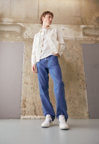 Veste en denim blanc, t-shirt blanc, jeans droits bleus et baskets blanches avec un design subtil, posés sur un fond en béton texturé.