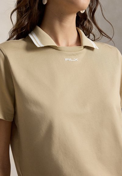 Žena nosící béžové polo tričko s bílými pruhy na límci a logem RLX na prsou, zobrazující detaily ramen a krku.