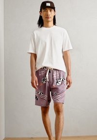 T-shirt en coton blanc, coupe ample. Shorts de bain violets clairs avec des motifs de parasols noirs et roses et de palmiers. Cordon de serrage à la taille.
