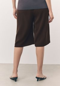 Culottes de satén marrón con un corte holgado, que terminan a mitad del muslo. Combinados con sandalias negras de tacón. Diseño simple, textura suave, sin estampados.