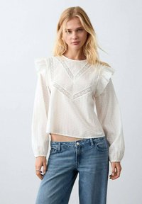 Bílá halenka s texturovanými puntíky, krajkovým detailem a volánkovými rukávy, doplněná světle modrými džínami.