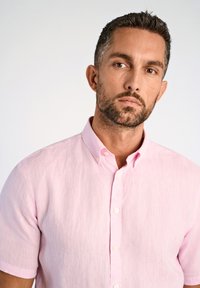 Camicia rosa chiaro a maniche corte con bottoni, realizzata in tessuto texturizzato, con colletto e dettagli intrecciati sottili, esposta su uno sfondo neutro.