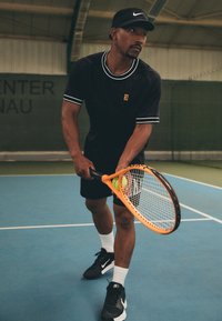 Černé sportovní tričko s bílým lemováním, černé šortky, černé boty Nike a kšiltovka. Drží oranžovou tenisovou raketu s tenisovými míčky na modrém kurtu.