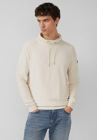 Kremowy pulower z wysokim kołnierzem, teksturowaną tkaniną, sznurkami i ściągaczami przy rękawach. W połączeniu z jasnoniebieskimi dżinsami.