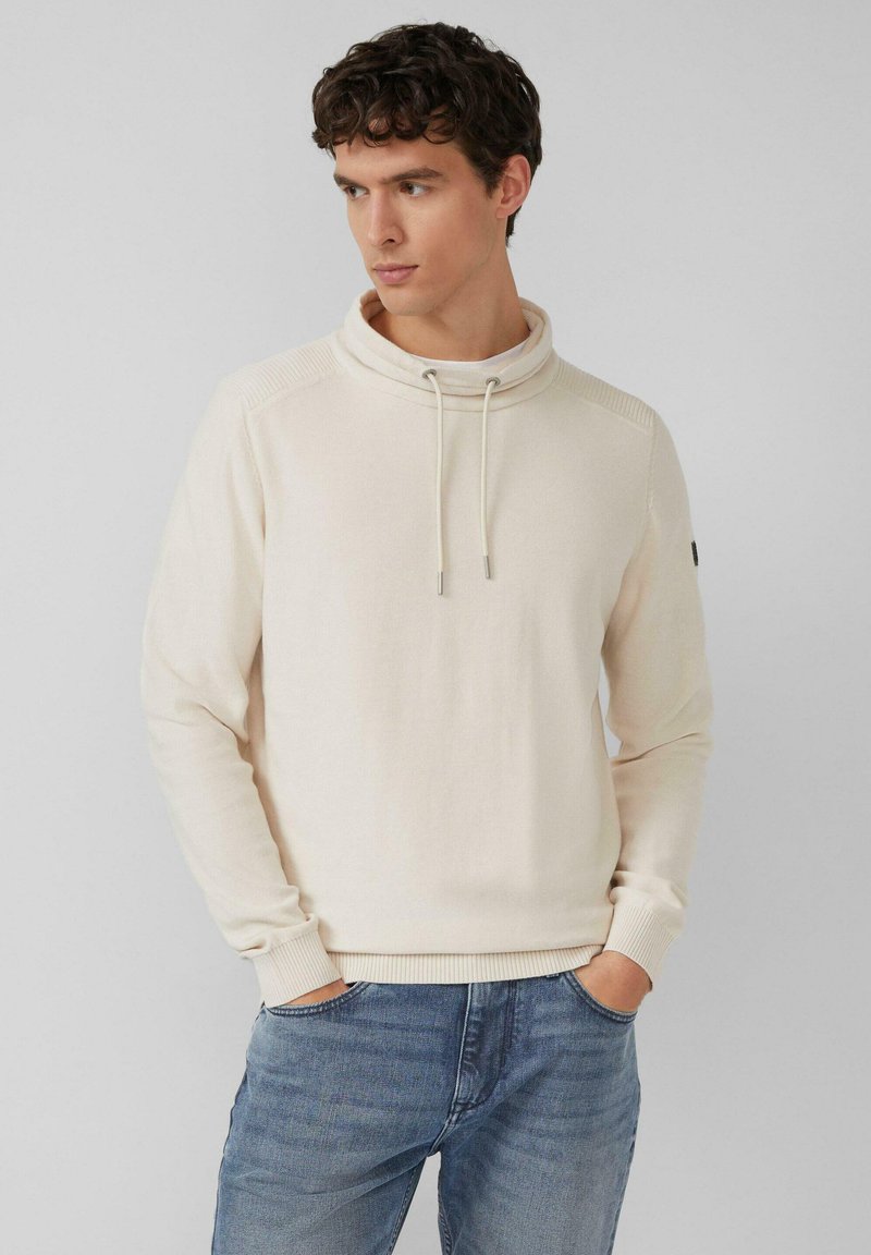 Kremowy pulower z wysokim kołnierzem, teksturowaną tkaniną, sznurkami i ściągaczami przy rękawach. W połączeniu z jasnoniebieskimi dżinsami.