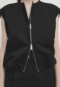 Haut noir sans manches avec deux fermetures éclair argentées partiellement fermées, porté sur un pantalon noir sur une personne à la peau claire.