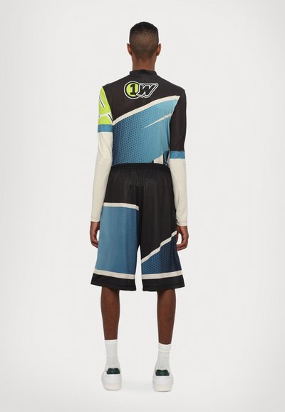 Sportsoutfit bestående af en langærmet top med blå og sorte geometriske mønstre, parret med sorte shorts og hvide sneakers.