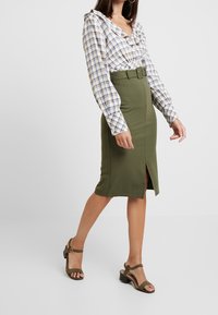 Olivgrüner Bleistiftrock mit einem Schlitz vorne und einem Gürtel, kombiniert mit einer weiß-blau karierten Bluse mit Rüschendetails und langen Ärmeln.