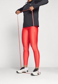 Sportliches Outfit mit einem schwarzen Langarmshirt und enganliegenden roten Leggings, dazu hält die Person ein rotes Widerstandsband. Sportliche Schuhe mit schwarz-weißem Design.