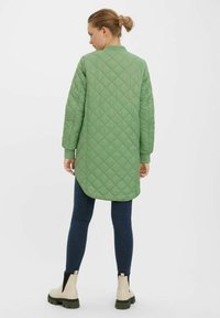 Cappotto verde trapuntato con scollo rotondo e maniche lunghe, caratterizzato da un orlo posteriore più lungo. Abbinato a jeans slim-fit e stivaletti beige alla caviglia.