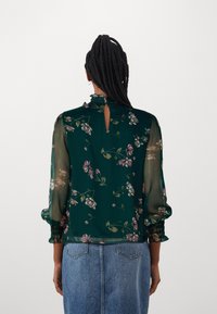 Blusa floreale verde con maniche lunghe trasparenti, scollatura arricciata, schiena a chiave e polsini elastici. Il motivo presenta fiori rosa e bianchi.