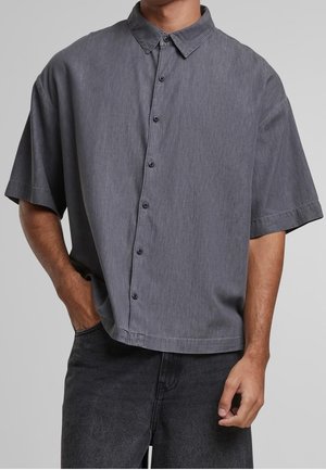 Mand iført en løstsiddende kortærmet mørkegrå skjorte med knapper og mørkegrå bukser, med den ene hånd i lommen.