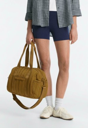 Persoon die marineblauwe shorts, witte sneakers, witte sokken en een geruit overhemd draagt en een mosterdkleurige doorgestikte reistas bij de handvatten vasthoudt.