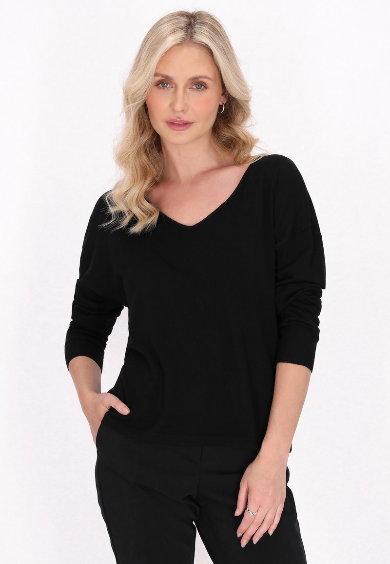 Pulover negru cu decolteu în V, dintr-un material moale și flexibil, având mâneci lungi și o croială lejeră. Modelul pozează cu o mână în buzunar, într-un stil minimalist.
