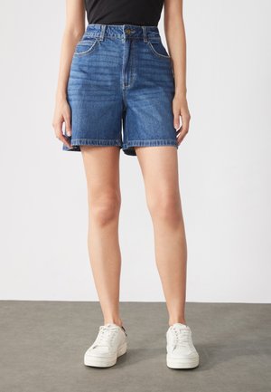 Denimshorts i mellanblått med hög midja, med sidofickor och fransad nederkant. I kombination med vita sneakers.