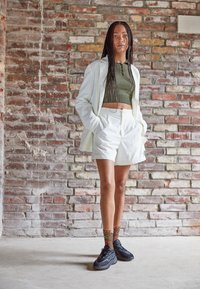 Jasnozielony, prążkowany top krótki, białe szorty w wysokim stanie oraz czarne sneakersy z kolorowymi, wzorzystymi skarpetkami. Tło wykonane z cegły.