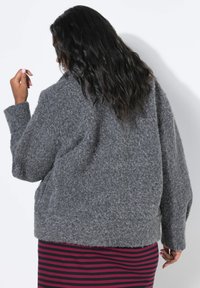 Maglione grigio, peloso e oversize con collo alto e spalle scese, abbinato a un vestito a righe con linee orizzontali rosse e nere.