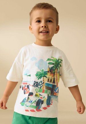 Jaunas berniukas dėvi baltą marškinėlį su spalvingais lenktyninių automobilių, palmės medžių ir pastato piešiniais, šypsodamasis priešais smėlio spalvos foną.