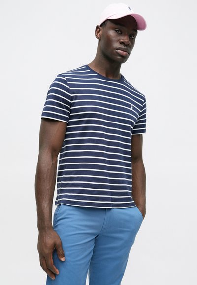 Μπλούζα με ναυτικές ρίγες σε σκούρο μπλε χρώμα και λευκές ρίγες, με κοντά μανίκια, στρογγυλή λαιμόκοψη και μικρό λογότυπο. Φοριέται με ανοιχτό μπλε παντελόνι και ροζ καπέλο.